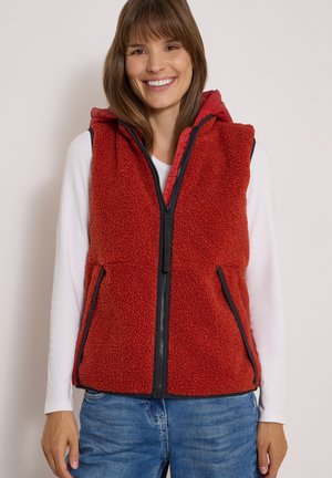 Femme souriante portant une veste sans manches en polaire rouge avec une fermeture éclair noire sur une chemise blanche à manches longues et un jean bleu.