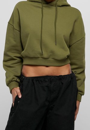 Osoba nosící olivově zelenou krátkou mikinu s kapucí a volné černé kalhoty s mašlí v pase, ruka v kapse, viditelný trup.