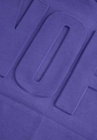 Fioletowy materiał z wypukłym wzorem w kształcie litery "V", gładką fakturą i widocznymi szwami wzdłuż krawędzi.