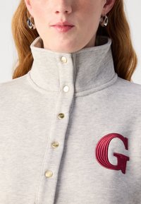 Gråkragad sweatshirt med guld snap-knappar, som har ett rött broderat bokstav "G" på vänster sida, tillverkad av ett mjukt, texturerat tyg.