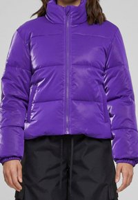 Fioletowa kurtka puchowa z wysokim kołnierzem, zapinana na zamek z przodu i elastycznymi mankietami. Posiada pikowaną strukturę i błyszczące wykończenie.