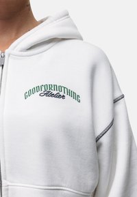 Witte hoodie met ritssluiting aan de voorkant, voorzien van groene geborduurde tekst "GOODFORNOTHING" en "Atelier", benadrukt met donkere stikdetails.