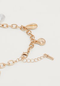 Gold chain bracelet featuring seashell and cowrie shell charms, with lobster clasp and brand tag. Smooth and polished texture.