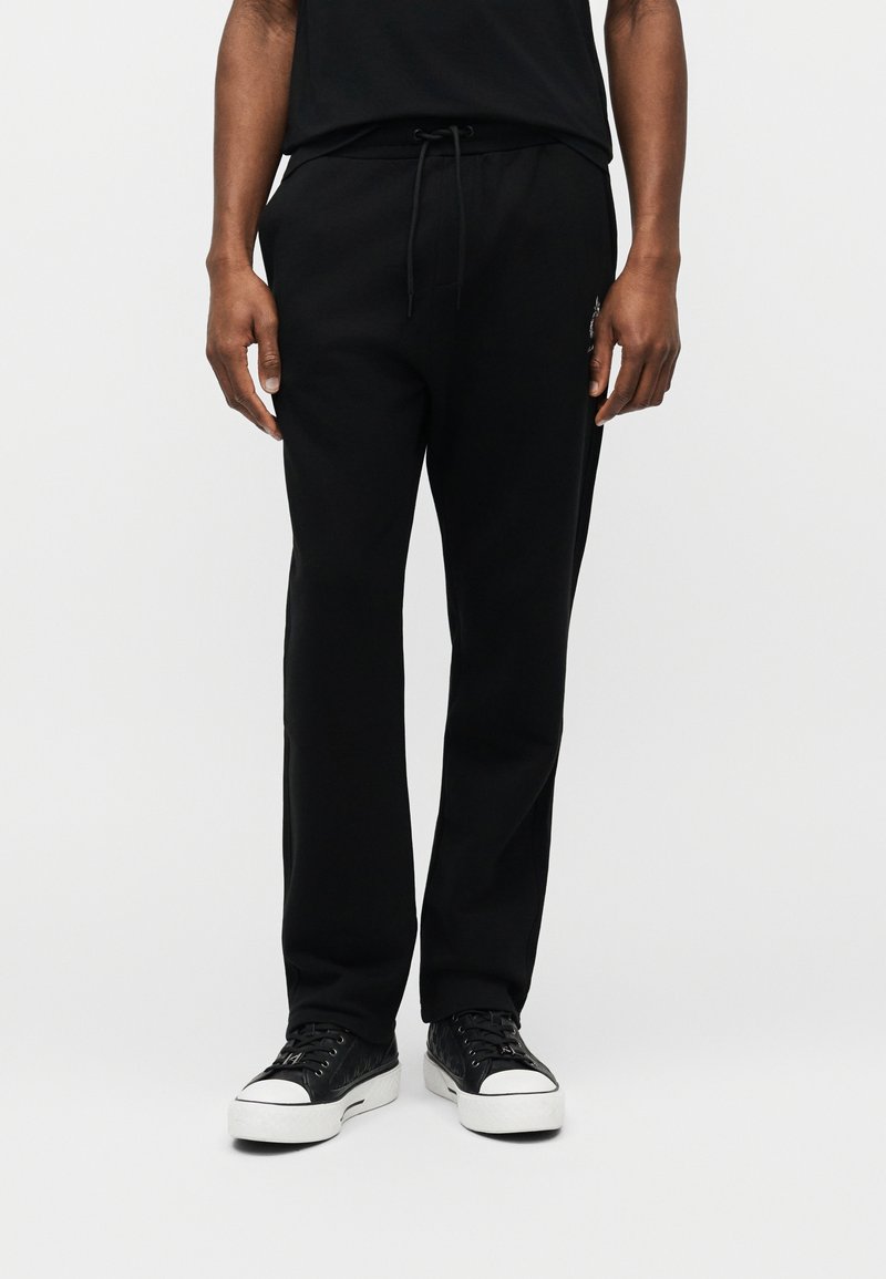 Άνδρας που φοράει μαύρο παντελόνι φόρμας με κορδόνι και μαύρα sneakers χαμηλής κοπής με λευκές σόλες, στέκεται μπροστά σε ανοιχτόχρωμο απλό φόντο.