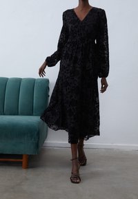 Czarna koronkowa sukienka w kwiaty z dekoltem w serek, długimi rękawami i talią podkreśloną paskiem. Zwiewna sylwetka; noszona z czarnymi sandałami na paski. W tle turkusowa kanapa.