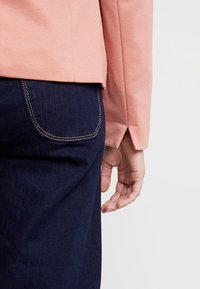 Vaaleanpunainen, teksturoitu pitkähihaisuus paita yhdistettynä tummansinisiin farkkuihin. Näkyvät käsi lepäävät lähellä paidan helmaa ja farkkujen taskusaumaa.