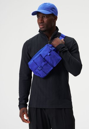 Blaues Nylon-Crossbody-Case mit verstellbarem Riemen und mehreren Fächern, getragen über einem schwarzen, strukturierten Langarmoberteil.
