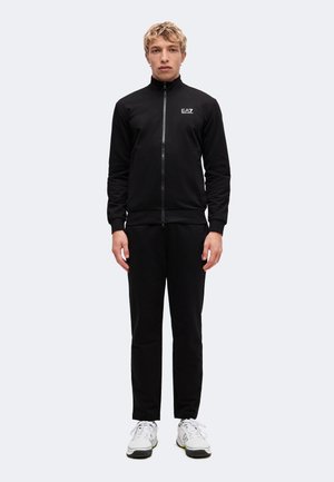 Homme blond debout portant une veste noire zippée et un pantalon avec des baskets blanches sur un fond clair uni.
