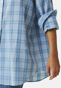 Modrá kostkovaná košile vyrobená z texturované látky, s knoflíkovou přední částí, ohrnutými rukávy a výraznými kostkovými vzory. Doplňuje ji detail s logem.