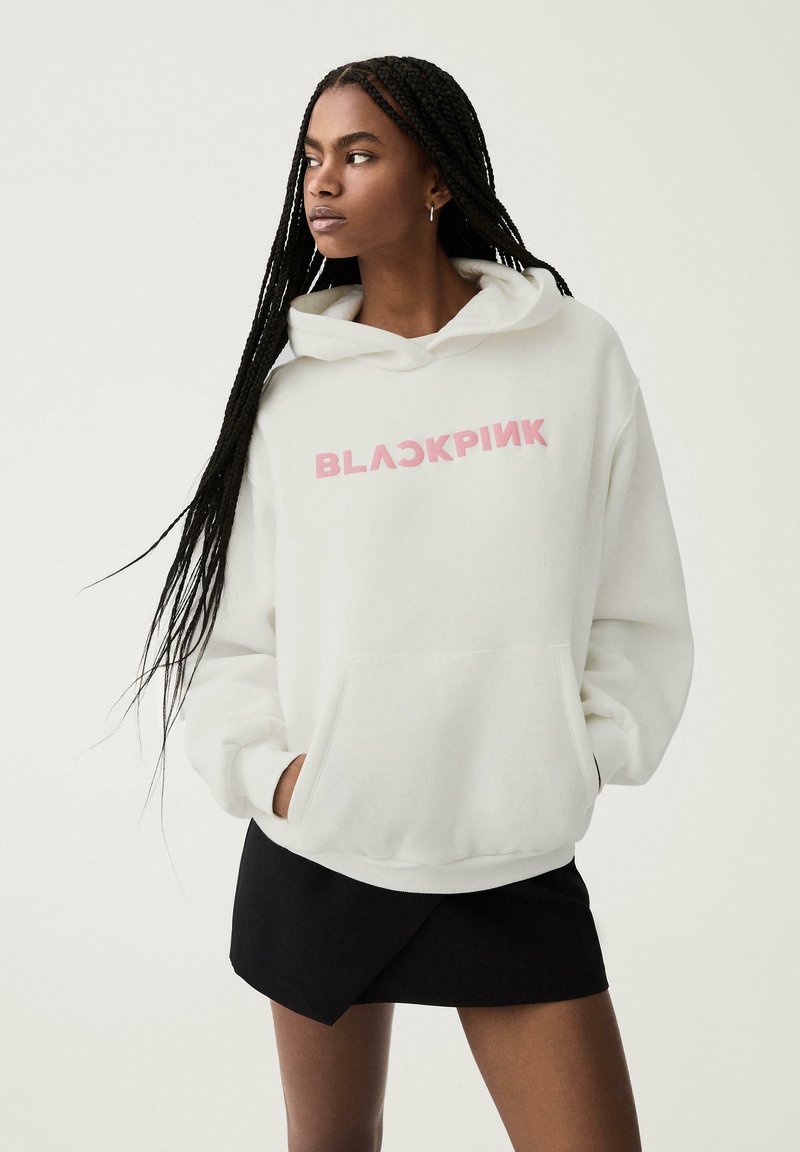 Sweatshirt à capuche blanche avec une poche frontale, arborant "BLACKPINK" en lettres roses. Porté avec une jupe noire, le tissu semble doux.