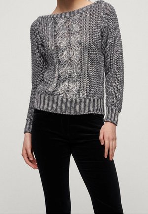Femme portant un pull en maille gris avec un motif en torsades central et des bordures côtelées aux poignets et à l'ourlet, associé à un pantalon noir.