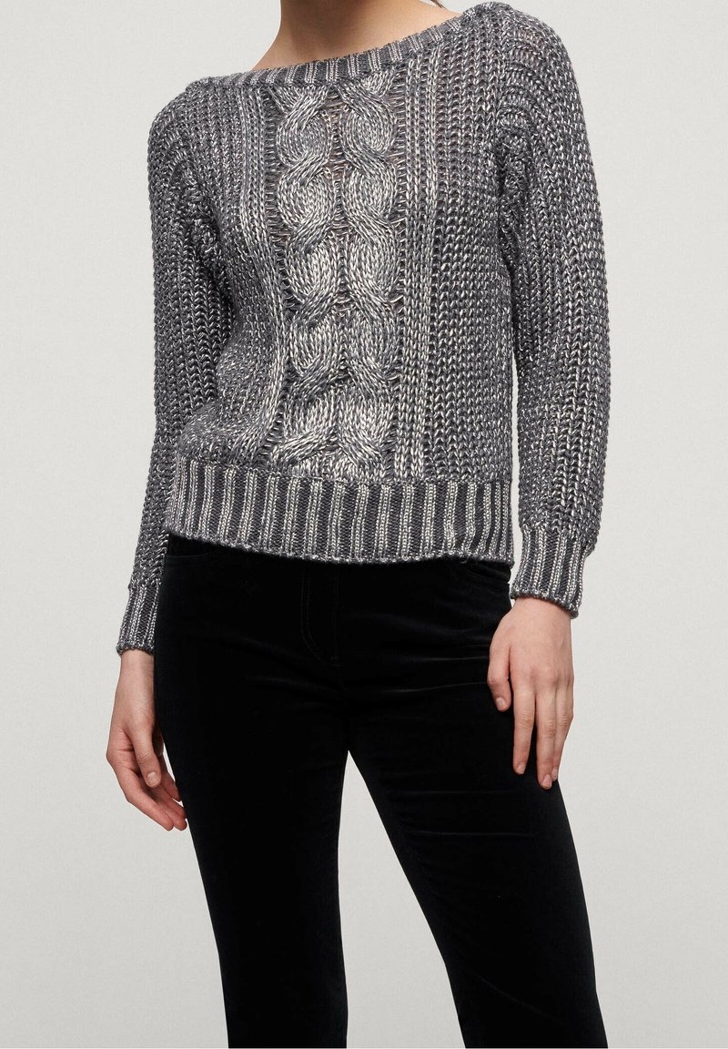 Femme portant un pull en maille gris avec un motif en torsades central et des bordures côtelées aux poignets et à l'ourlet, associé à un pantalon noir.