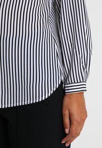 Blouse à rayures noir et blanc avec un ourlet arrondi, des poignets boutonnés et un tissu léger. Associée à un pantalon noir, montrant une main sur le côté.