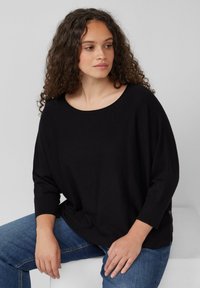 Zwarte, losvallende longsleeve top met een brede halslijn. Zachte textuur, gedragen met blauwe jeans. Opvallend detail aan de zijkant van de zoom.