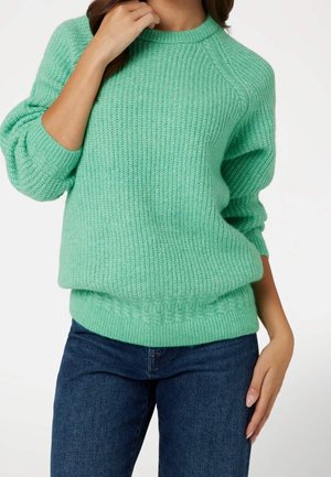 Femme portant un pull en maille vert clair et un jean bleu, une main touchant son cou et l'autre reposant à ses côtés.