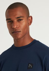 Navyblaue Baumwoll-T-Shirt mit einem kleinen schwarzen Quadrat-Patch mit einem weißen Logo, Rundhalsausschnitt und kurzen Ärmeln. Glatte Textur.