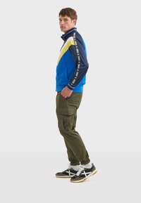Blå zipjacka med gula och vita detaljer samt logotape, tillsammans med olivgröna cargobyxor och svarta sneakers med vita ränder.
