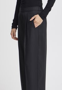 Zwarte, op maat gemaakte broek met een hoge taille, geplooid ontwerp en zijzakken. Gladde stof met een lichte glans en een aangesloten tailleband.