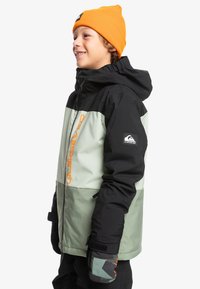 Svart och mintgrön isolerad jacka med huva, med en orange logotyp och detaljer. Modellen har på sig en matchande orange mössa och mönstrade handskar.