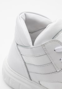 Detailní záběr na bílé vysoké tenisky s polstrovaným kotníkovým límcem, detaily šití a částí šněrování na bílém pozadí.