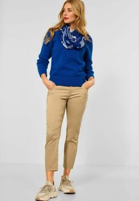 Blauwe gebreide trui, beige cropped broek en beige sneakers met textuuraccenten. Blauwe patroon sjaal wikkelt om de nek.