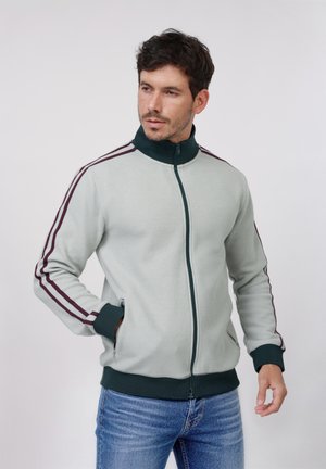 Uomo che indossa una giacca grigia chiaro con cerniera e rifiniture verde scuro e strisce bordeaux sulle maniche, abbinata a jeans blu, in piedi davanti a uno sfondo neutro.