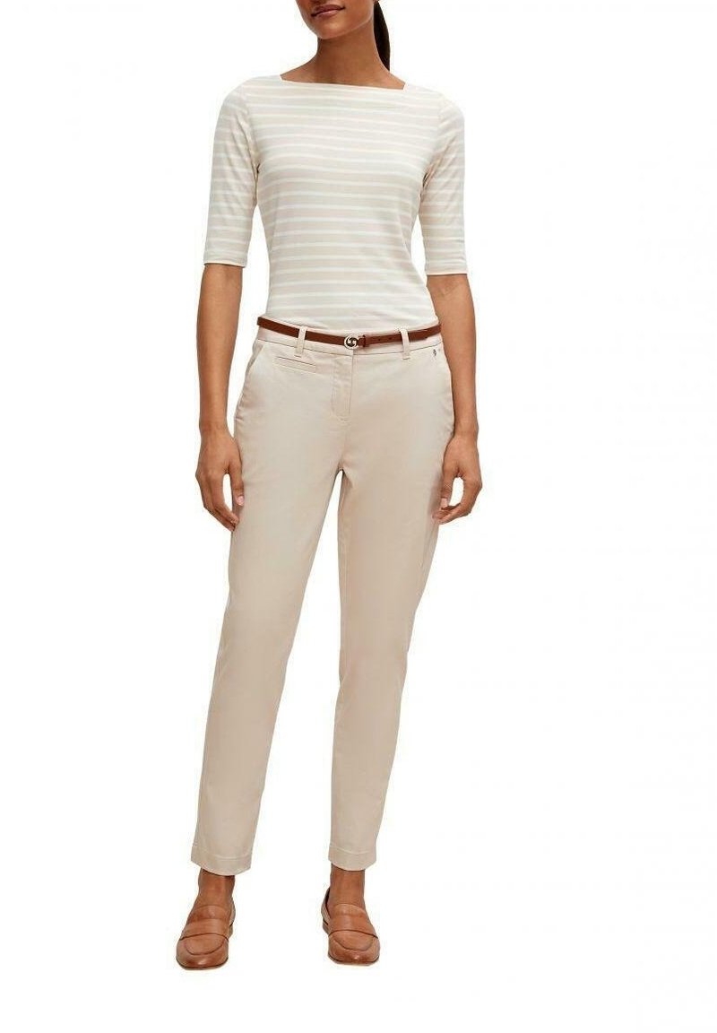 Gestreiftes, langärmliges Oberteil in Weiß und hellbeige; beiges Slim-Fit-Hose mit einem braunen Gürtel. Braune Slipper mit flachem Absatz.