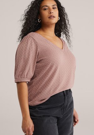 Göndör hajú nő ezüst karika fülbevalóval, rózsaszín texturált V-nyakú felső puffos ujjal és magas derekú fekete nadrággal.