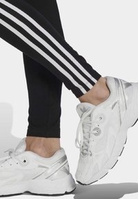 White athletic shoes with mesh and synthetic textures, featuring silver accents and a black rubber sole, paired with black leggings with white stripes.