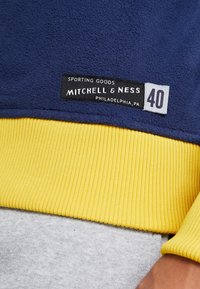 Tissu polaire bleu avec une étiquette Mitchell & Ness Philadelphia PA noire et blanche et un ourlet côtelé jaune numéroté 40, porté sur un pantalon gris.