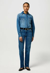 Mujer de pie con camisa de botones de denim azul y jeans, con cinturón negro y zapatos negros, posando frente a un fondo claro y liso.