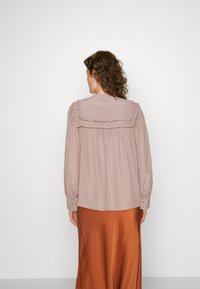 Långärmad, ljus mauveblus med volangdetaljer längs kragen och axlarna, kombinerad med en flödande kjol i rostfärg.