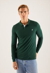 Mørkegrønn langermet polo skjorte med hvit krage, med en liten hvit logo på brystet og en tilpasset design. Brukt sammen med lyseblå jeans.