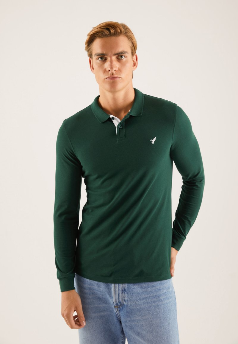 Mørkegrønn langermet polo skjorte med hvit krage, med en liten hvit logo på brystet og en tilpasset design. Brukt sammen med lyseblå jeans.