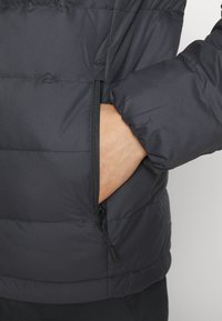 Dettaglio di un giubbotto puffo nero che mostra una tasca laterale con zip. Il tessuto appare liscio con una texture trapuntata, con maniche a polsino.