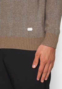 Brauner Pullover mit gerippten Bündchen und Saum, der eine dezente Textur aufweist und ein gesticktes Markenlabel in der unteren Ecke hat. Schwarze Hose sichtbar.