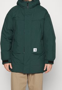 Man die een donkergroen gewatteerd winterjack met voorzakken en een capuchon draagt, gecombineerd met beige broek, staand tegen een effen achtergrond.