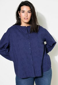 Camicia blu navy con bottoni caratterizzata da un motivo floreale testurizzato, scollatura rotonda e vestibilità comoda, abbinata a jeans in denim blu.