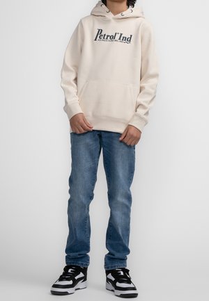 Barn iført en beige Petrol Ind hættetrøje, blå jeans og sort-hvide sneakers, stående foran en ensfarvet baggrund.