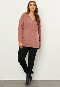Maglione rosa a maglia con scollo a V e texture a zigzag, abbinato a jeans neri slim-fit e stivaletti neri con tacco.