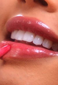 Close-up van glanzende, roze lippen met een glanzende afwerking, waarbij witte tanden te zien zijn. Een hand brengt lipstick aan met een felroze applicator.