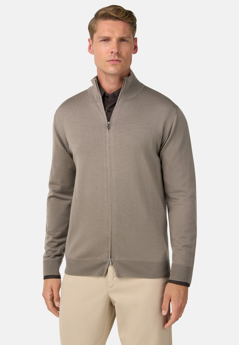 Bežna zip-up jopica z teksturiranim vzorcem, rebrastimi manšetami in robom, v kombinaciji s temno srajco s ovratnikom. Nosijo jo model v nevtralnem okolju.