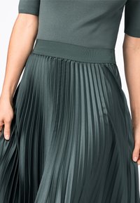 Gonna midi plissettata di colore verde scuro, con una cintura liscia e pieghe verticali, realizzata in un tessuto leggero e fluido.