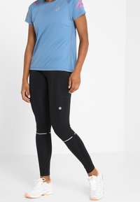 Camiseta deportiva de manga corta azul claro con detalles en rosa, combinada con leggings negros con detalles reflectantes y un logotipo. Zapatillas blancas.