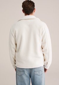 Jachetă albă din fâș cu o suprafață texturată, guler supradimensionat și o croială lejeră, asortată cu blugi din denim de culoare bleu deschis.