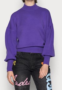 Maglione a collo alto di colore viola, con maniche ampie e polsini a coste. Indossato con jeans in denim scuro che presentano accenti di testo colorati.