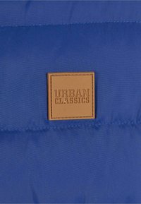 Sezione di giacca imbottita blu royal con una toppa quadrata in pelle beige impresso con "URBAN CLASSICS." Texture liscia con cuciture visibili.