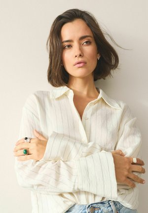 Weißes Hemd mit dünnen vertikalen Streifen, hellblaue Jeansshorts, mehrere Ringe, darunter ein mit grünem Stein, und ein einfacher Ohrstecker.