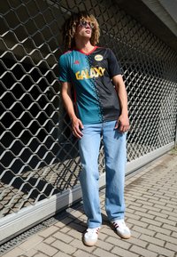 Camiseta de Adidas del LA Galaxy en azul verdoso y negro con un patrón de estrellas, acentos rojos, combinada con jeans de un azul claro y zapatillas blancas con rayas rojas.