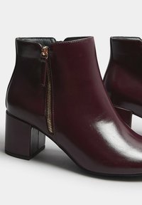 Botas de tobillo burdeos brillantes con tacones de bloque y cierres de cremallera dorados laterales sobre un fondo blanco liso.